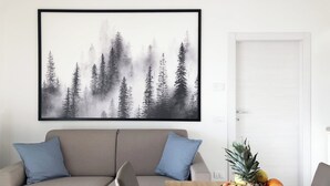 A living room with a beige sofa, two blue cushions, a wooden dining table with a fruit bowl, and a framed forest landscape on the wall. - S02 - 4 PAX – TERRACE (LAVARONE)