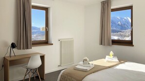 A hotel room with a bed, a desk, a chair, and two windows offering a mountain view. - 002 - 4 PAX – TERRACE (LAVARONE)