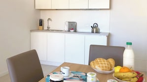 Une cuisine moderne avec des meubles blancs, un évier et un grille-pain. On y trouve une table à manger avec un saladier de fruits, une bouteille de lait et un pot de confiture.
