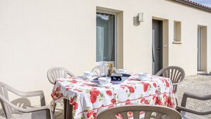 A table set outdoors with a floral tablecloth, surrounded by chairs, against a building with a tiled roof and a window.