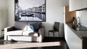 A modern living room with a sofa and a chair, a kitchen area with a sink and cabinets, and a large cityscape painting on the wall. - 104 – 6 PAX TERRACE (LAVARONE)