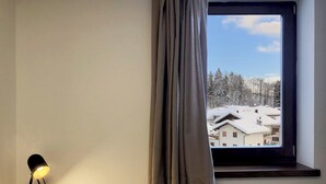 A room with a window overlooking a snowy village, a lamp on a wooden table, and a radiator below the window. - 104 – 6 PAX TERRACE (LAVARONE)