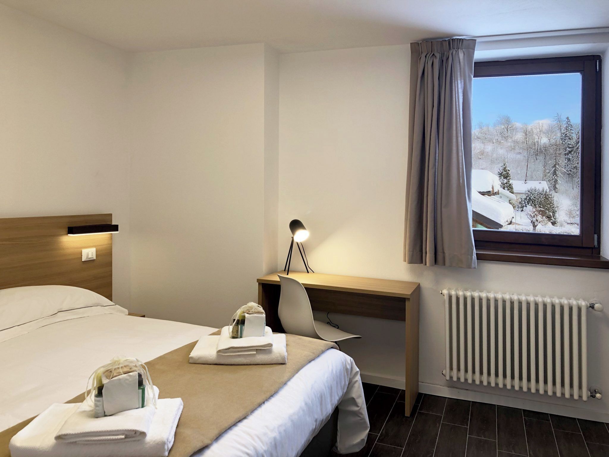 A hotel room with a bed, a desk with a lamp, a chair, a window with curtains, and a radiator.