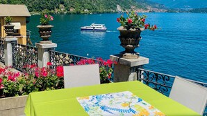 A terrace with a table, chairs, and potted plants overlooking a lake with a boat and mountains in the background.