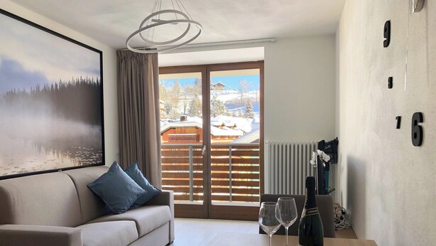A modern living room with a sofa, a dining table with a bottle of wine, glasses, and food. A large window offers a view of a snowy landscape. - 2+2 apartment with large panoramic terrace (202) (LAVARONE)