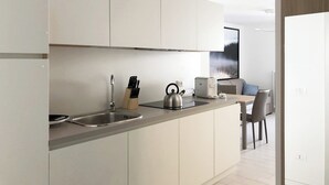 Una cocina moderna con armarios blancos, un lavamanos de acero inoxidable y una tetera. Se ve un comedor con una mesa y sillas al fondo.
