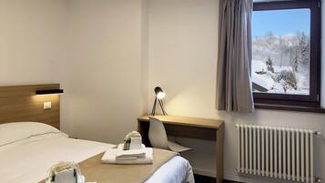 A hotel room with a bed, a desk with a lamp, a chair, a window with curtains, and a radiator.