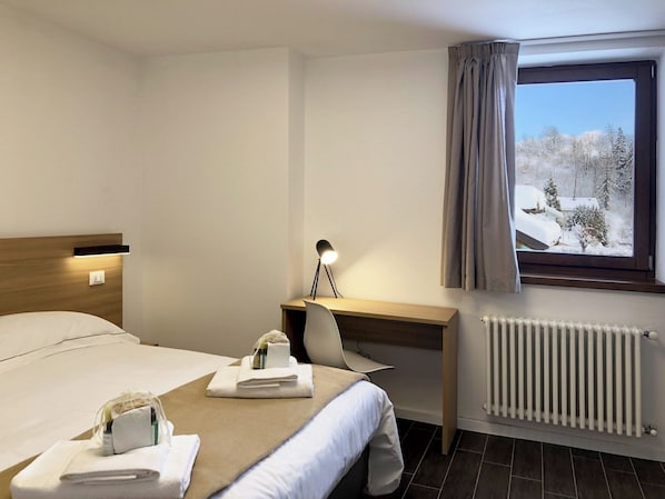 A hotel room with a bed, a desk with a lamp, a chair, a window with curtains, and a radiator.