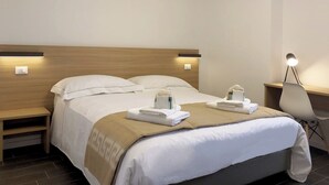 Un lit bien fait, avec du linge de lit blanc et un plaid beige, une table de chevet avec une lampe, et une chaise près d’un bureau, également avec une lampe.