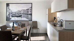 Une cuisine moderne équipée d’un grille-pain encastré, d’une bouilloire et d’un évier. On y trouve un coin repas avec une table dressée pour deux personnes, ainsi qu’une photographie en noir et blanc encadrée accrochée au mur.