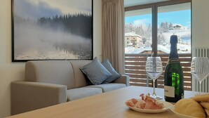Un salon moderne avec un canapé, un tableau représentant un paysage enneigé, une table à manger avec une bouteille de vin, des verres et du pain.