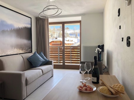 A modern living room with a sofa, a dining table with wine glasses and a bottle, and a view of a snowy landscape through the sliding doors.