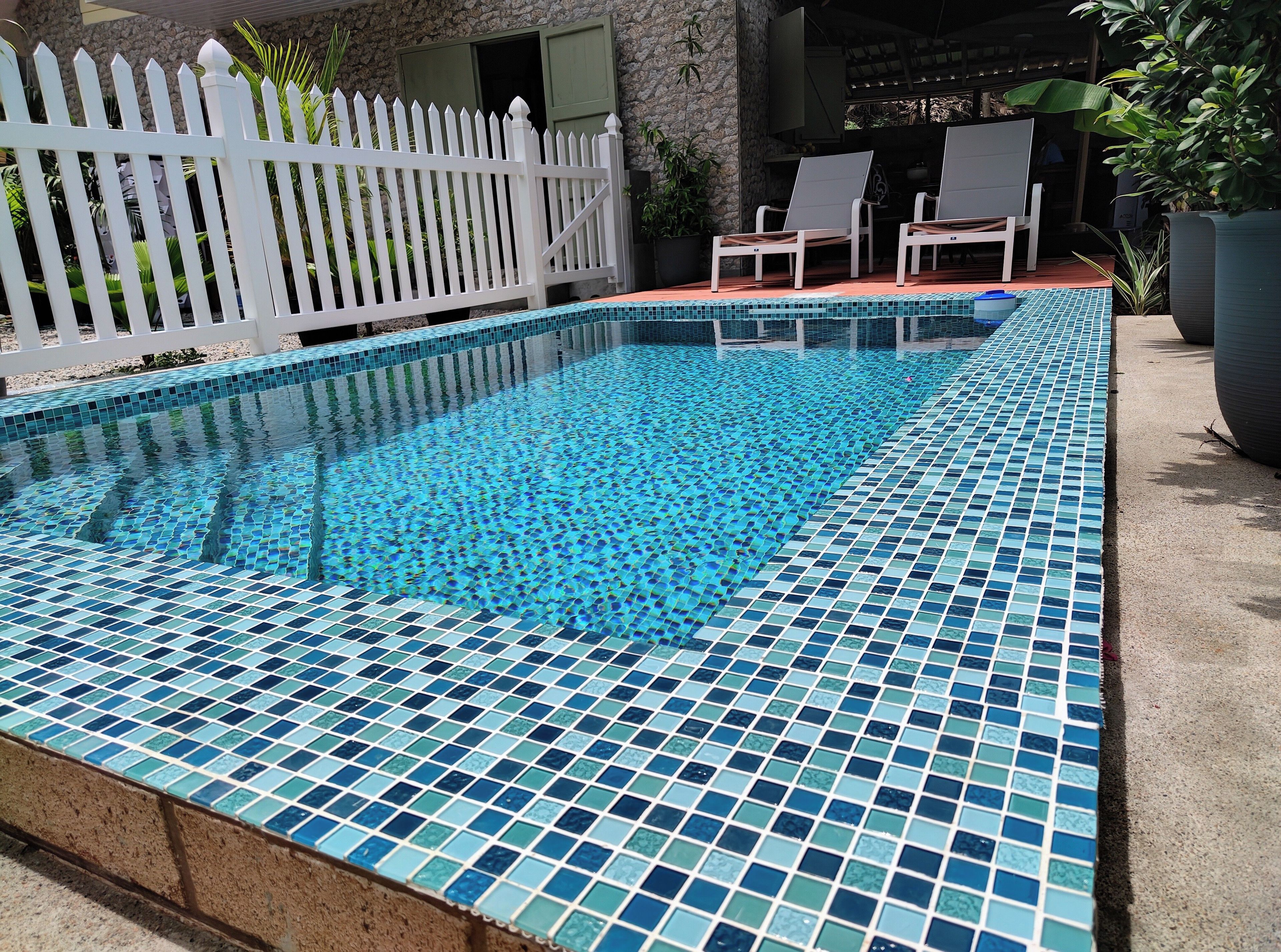 Una piscina rectangular con mosaicos azules, rodeada por una cerca blanca de piquete y dos sillas de descanso.