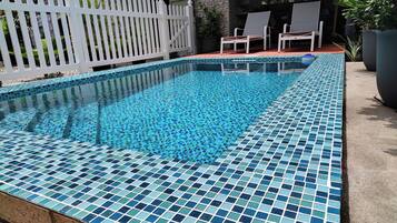 Una piscina rectangular con mosaicos azules, rodeada por una cerca blanca de piquete y dos sillas de descanso.