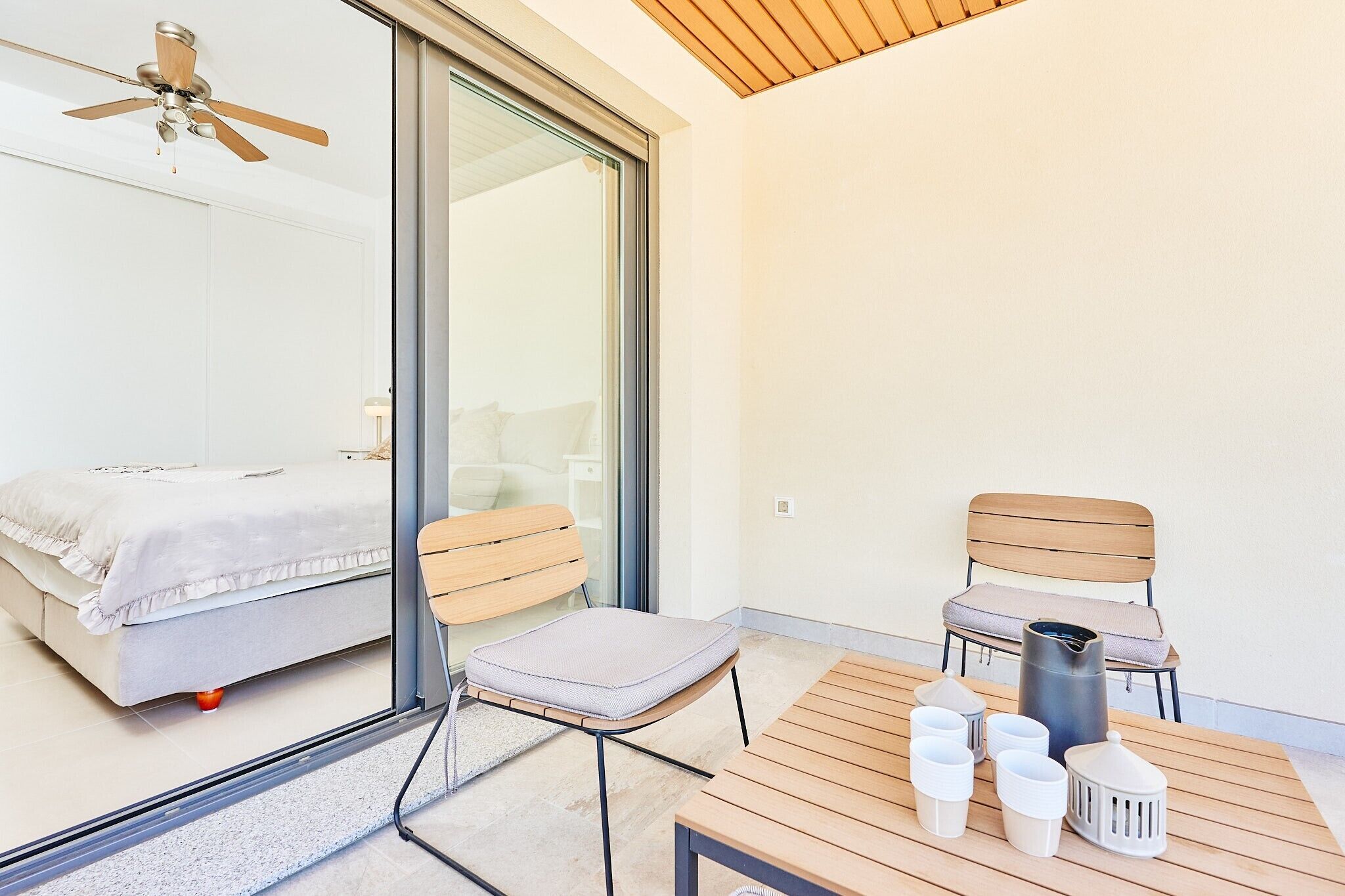 A balcony with a bed, two chairs, a table with cups and a container, and a ceiling fan.