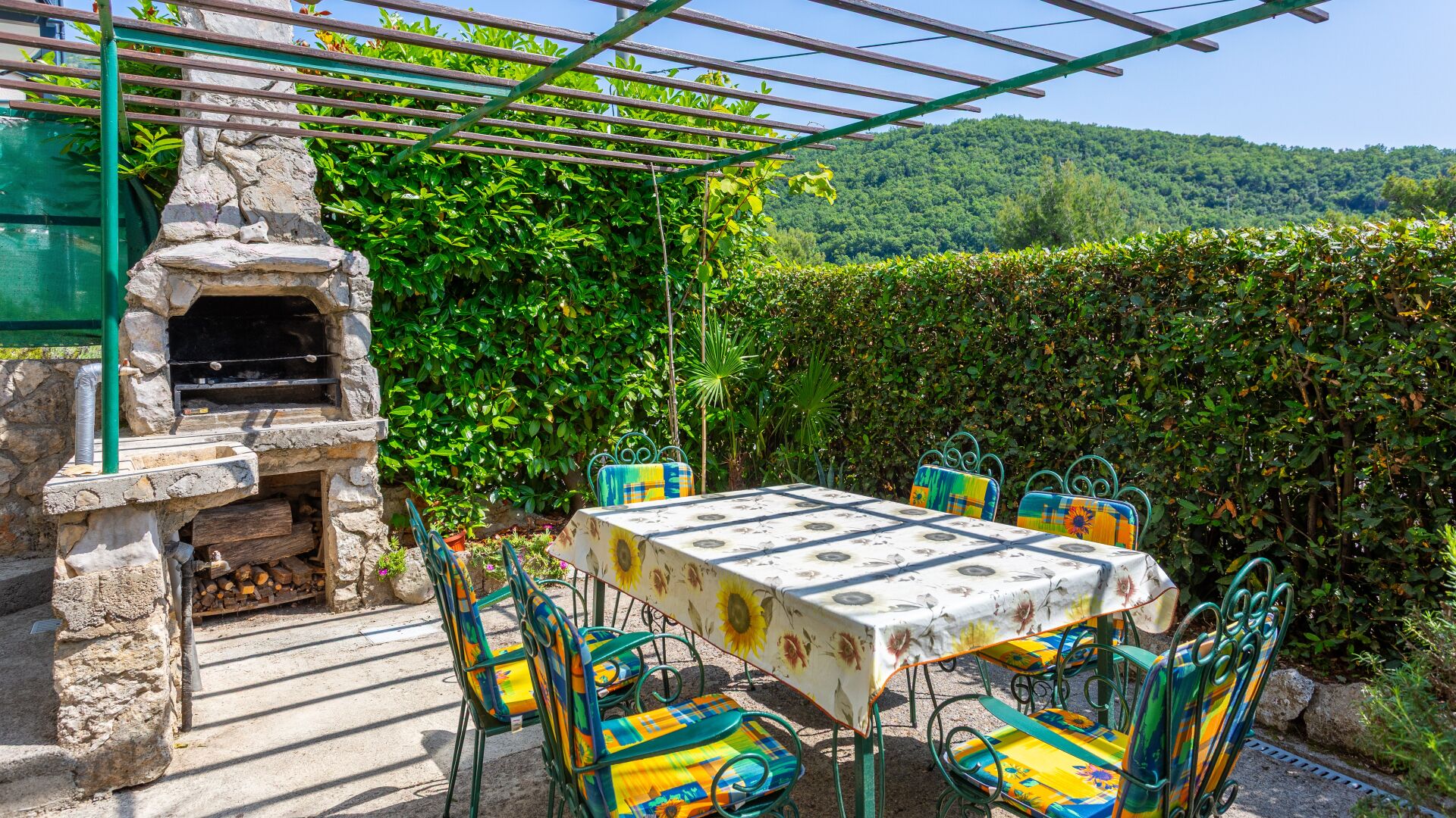 Une terrasse extérieure avec un barbecue en pierre, une table recouverte d’une nappe à motifs floraux et des chaises colorées.