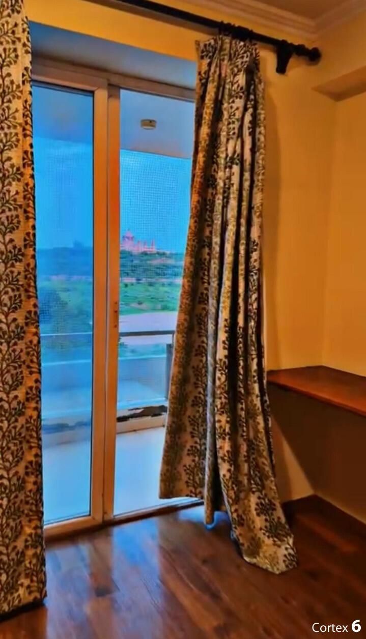 A room with a sliding glass door leading to a balcony, patterned curtains, and a wooden floor.