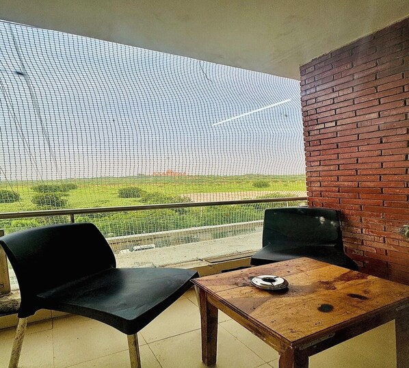 Un balcon avec deux chaises noires et une table en bois, donnant sur un paysage verdoyant, avec un mur de briques sur un côté.