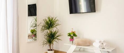 A modern bathroom with a wooden floor, a white vanity, a potted plant, a mirror, and a flat-screen TV.