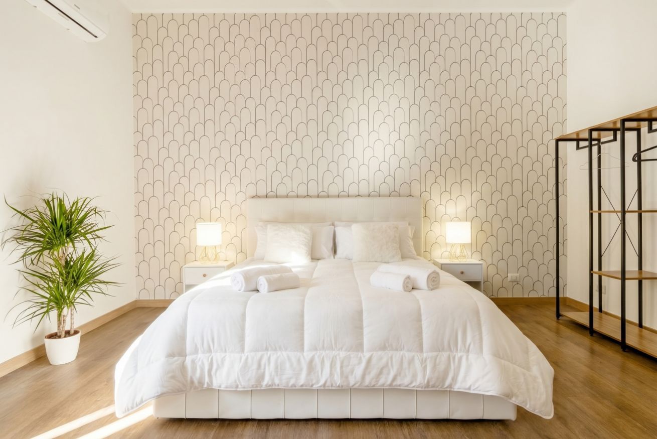Un lit bien fait, recouvert d’une couette blanche, deux tables de chevet avec des lampes, une plante en pot et un mur orné d’un motif.
