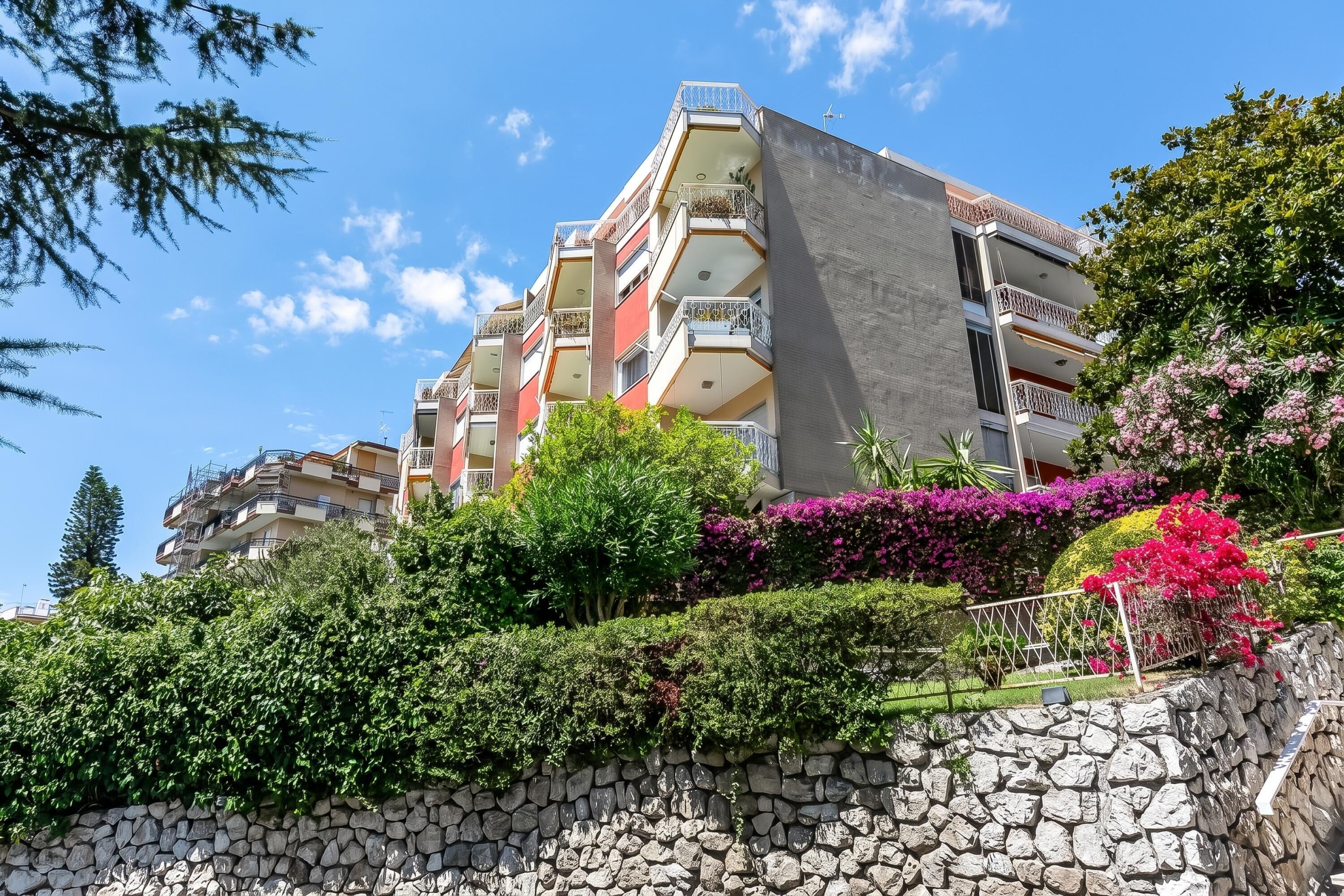 A multi-story building with balconies, surrounded by lush greenery and flowering plants.