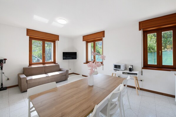 A modern living room with a wooden dining table, white chairs, a beige sofa, a flat-screen TV, and a small kitchen area with a microwave.