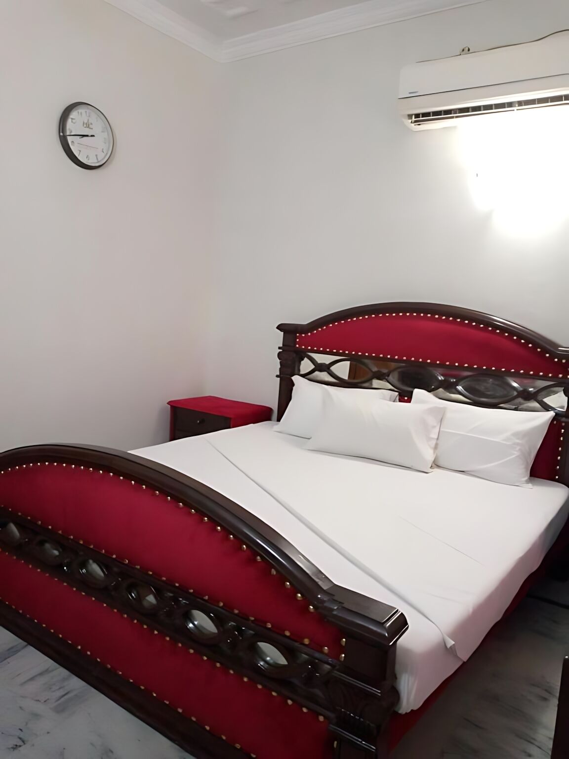 A bedroom with a red upholstered headboard, white bedding, a bedside table, and a wall clock.