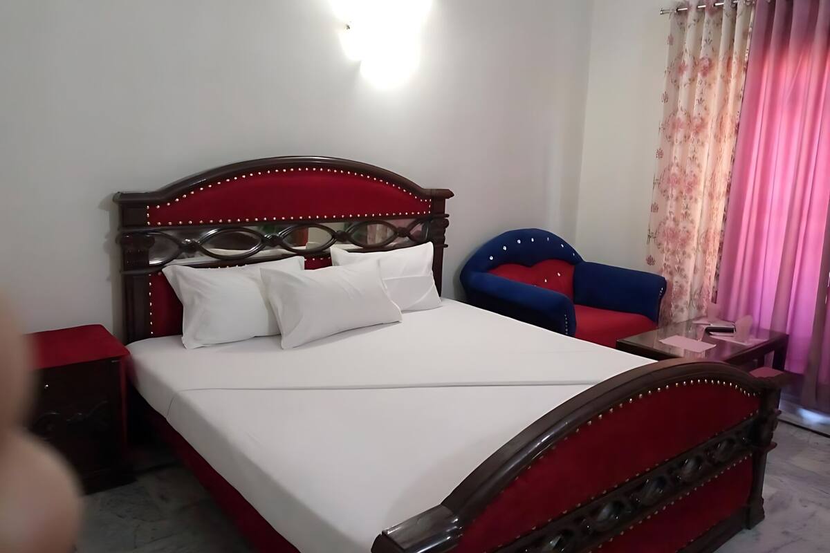 Un dormitorio con una cama tapizada de rojo, cabecera de madera, almohadas blancas, una mesita de noche y una silla azul.