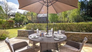 Une table de jardin ronde, entourée de chaises en osier et surmontée d’un grand parasol, est installée dans un jardin agrémenté d’un mur de pierre et d’un bâtiment visible au loin.