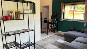 A modern living room with a grey sofa, a black metal shelf with books and glasses, a dining area with a table and chairs, and a window with a view of a building outside.