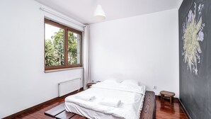Een slaapkamer met een bed, een raam, een radiator, een klein houten krukje en een muur met een groot abstract schilderij.