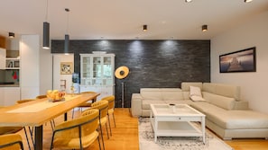 Een moderne woonkamer met een bank, een eettafel met stoelen, een keukenhoek en een groot raam.