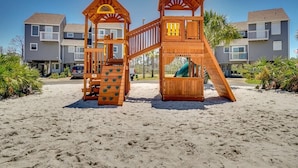 Een houten speeltoestel op een zandstrand, met een woongebouw op de achtergrond.