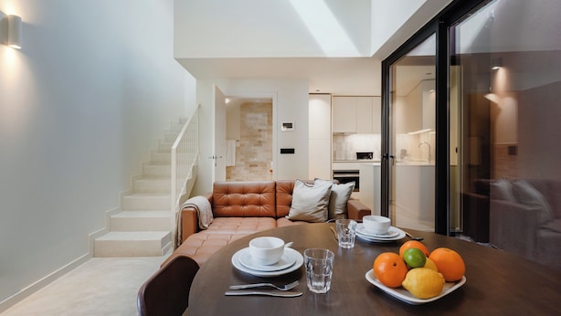 Un comedor con una mesa dispuesta para dos, una escalera, una sala de estar con un sofá de cuero marrón y una cocina al fondo.