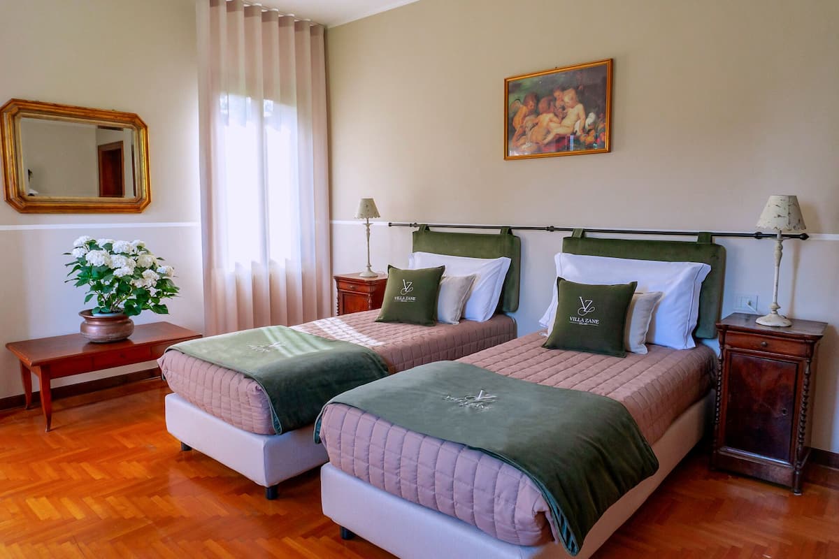 A hotel room with two beds, a painting on the wall, a mirror, and a window with curtains.