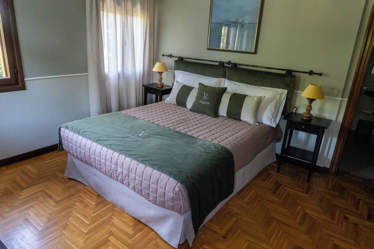 A neatly made bed with a quilted bedspread, two bedside tables with lamps, a headboard, and a painting above the bed.