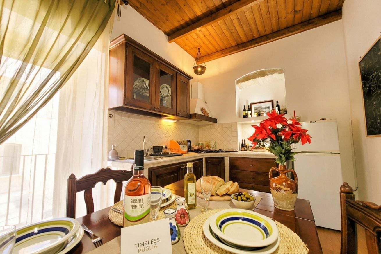 Un comedor con una mesa de madera dispuesta para una comida, una cocina al fondo y un jarrón con flores rojas sobre la mesa.