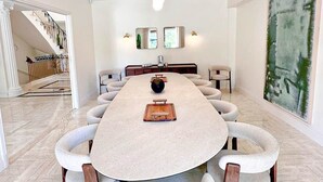 Un vaste espace dédié à la salle à manger, avec une longue table et de nombreuses chaises, un escalier en arrière-plan et une grande peinture abstraite accrochée au mur.