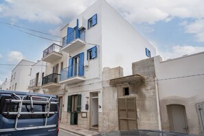 A multi-story building with balconies, a car parked in front, and a stone wall.