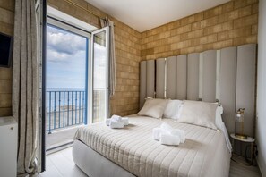 A hotel room with a large bed, a view of the sea through a balcony, and a modern design with a brick wall and a headboard with vertical panels.