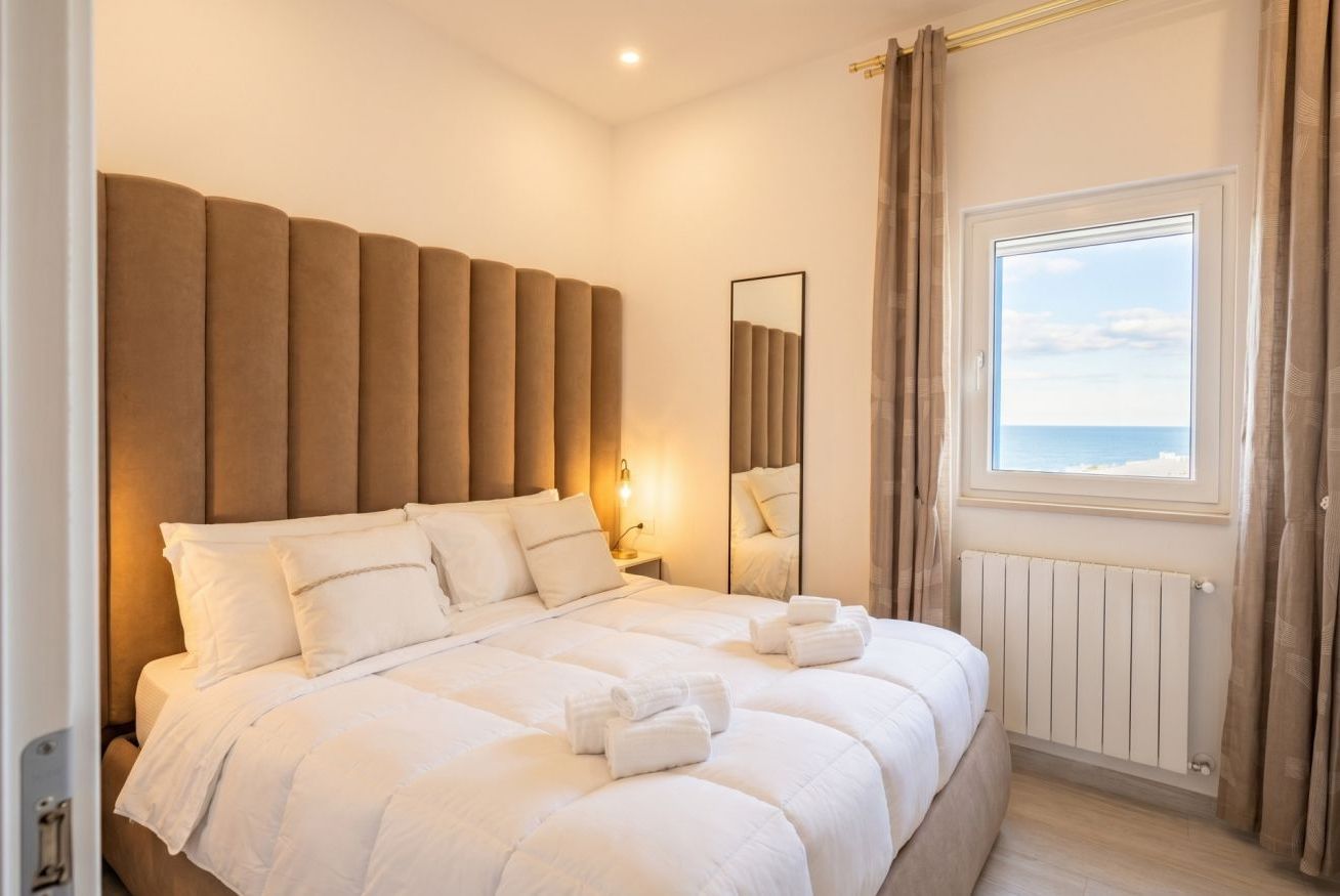 A hotel room with a large bed, a nightstand, a mirror, a window with curtains, and a view of the sea.