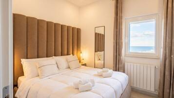 A hotel room with a large bed, a nightstand, a mirror, a window with curtains, and a view of the sea.