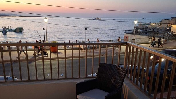 Un balcón con mobiliario de mimbre y una mesa, con vista a un muelle con gente y barcos.