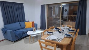 Una sala de estar con un sofá azul, una mesita blanca pequeña y una mesa de comedor preparada para cuatro con sillas de madera.