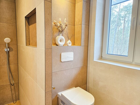 A modern bathroom with a shower, toilet, and a window providing a view of trees.