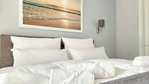 Uma cama bem arrumada com lençóis e almofadas brancas, um quadro com uma cena de praia na parede e uma luminária fixada na parede.