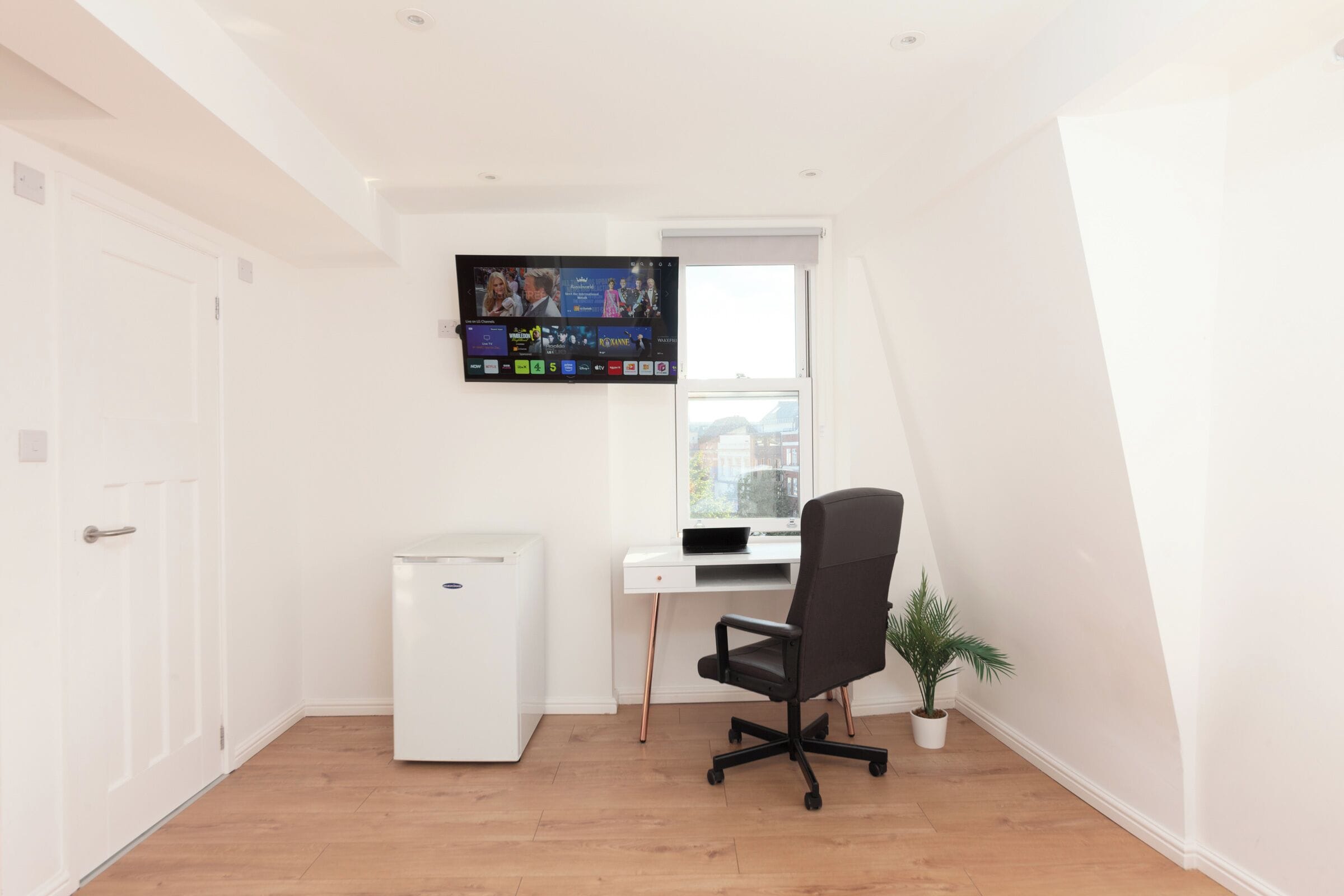A small, bright room with a white door, a white desk with a computer, a black office chair, a small white refrigerator, and a TV mounted on the wall.