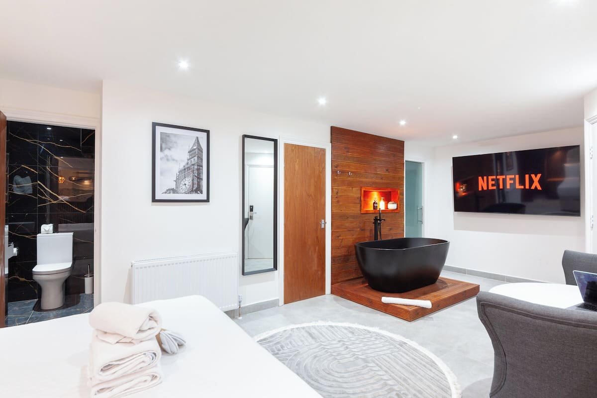 A modern hotel room with a large flat-screen TV, a black bowl-shaped bathtub, a wooden accent wall, and a framed picture of a clock tower.