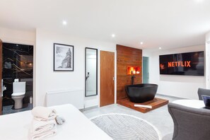 A modern hotel room with a large flat-screen TV, a black bowl-shaped bathtub, a wooden accent wall, and a framed picture of a clock tower. - Villas Apart Hotel (London)