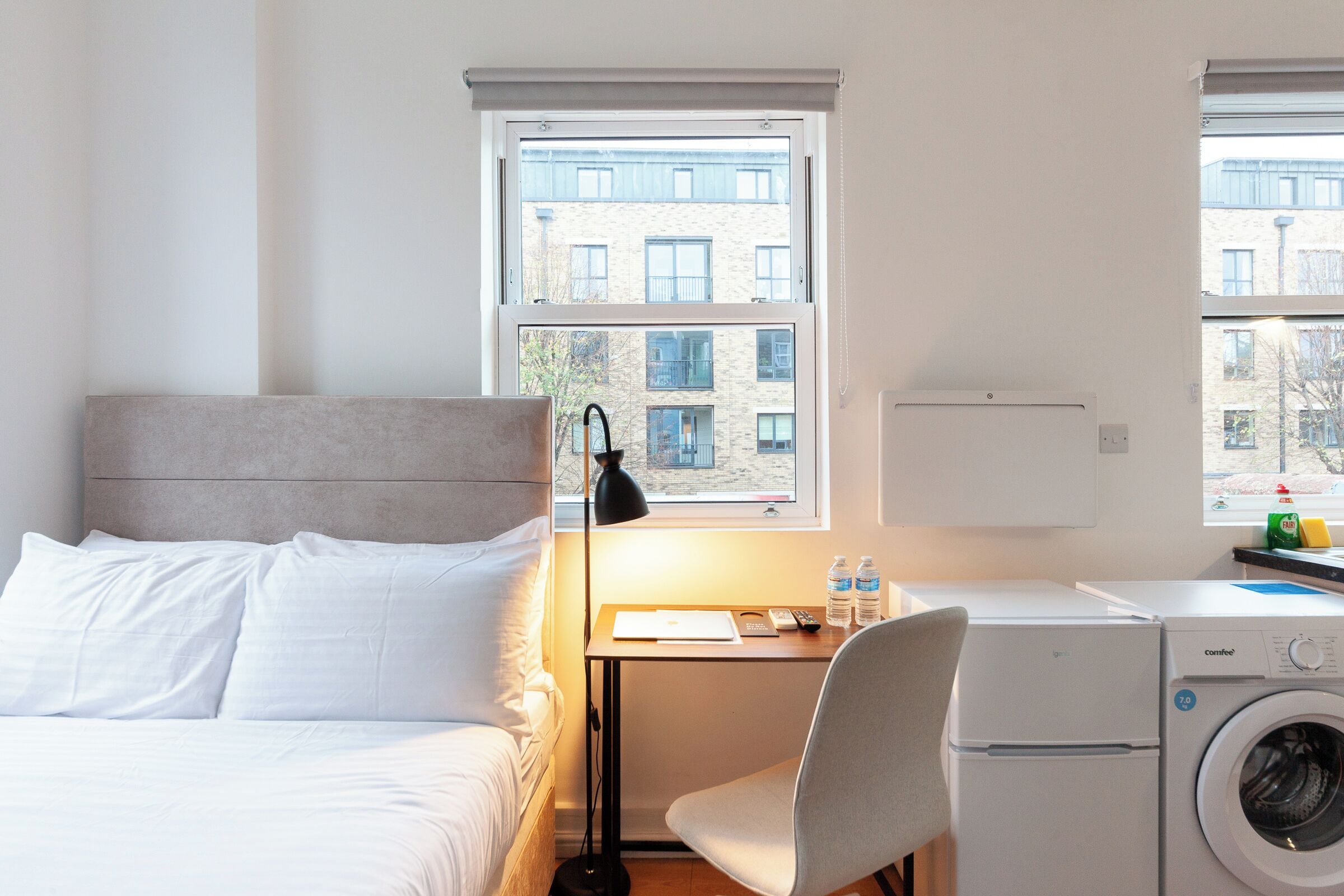 A bedroom with a bed, a desk, a chair, a washing machine, and windows with a view of buildings.